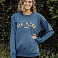 Person wearing a blue sweatshirt with 'Raising Warriors' text, standing against a green leafy background