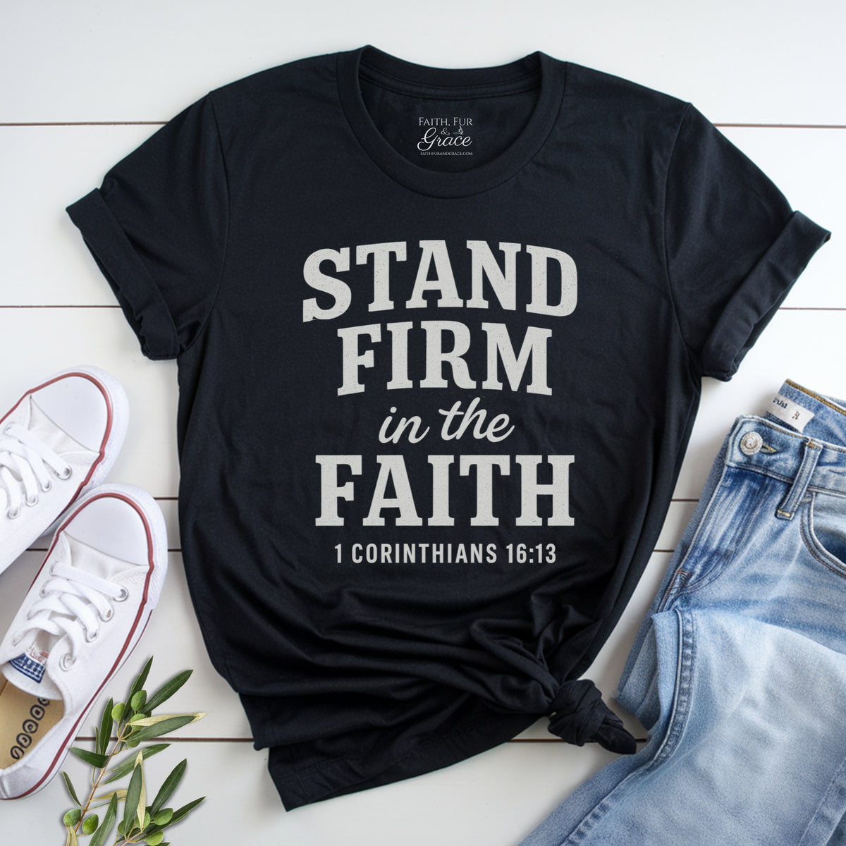Black t-shirt with 'Stand Firm in the Faith' text on a white wooden surface with shoes and jeans.