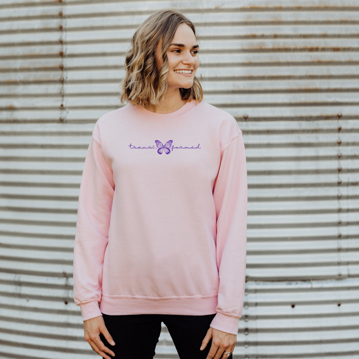 Woman wearing a pink sweatshirt with text and a butterfly graphic against a corrugated metal background