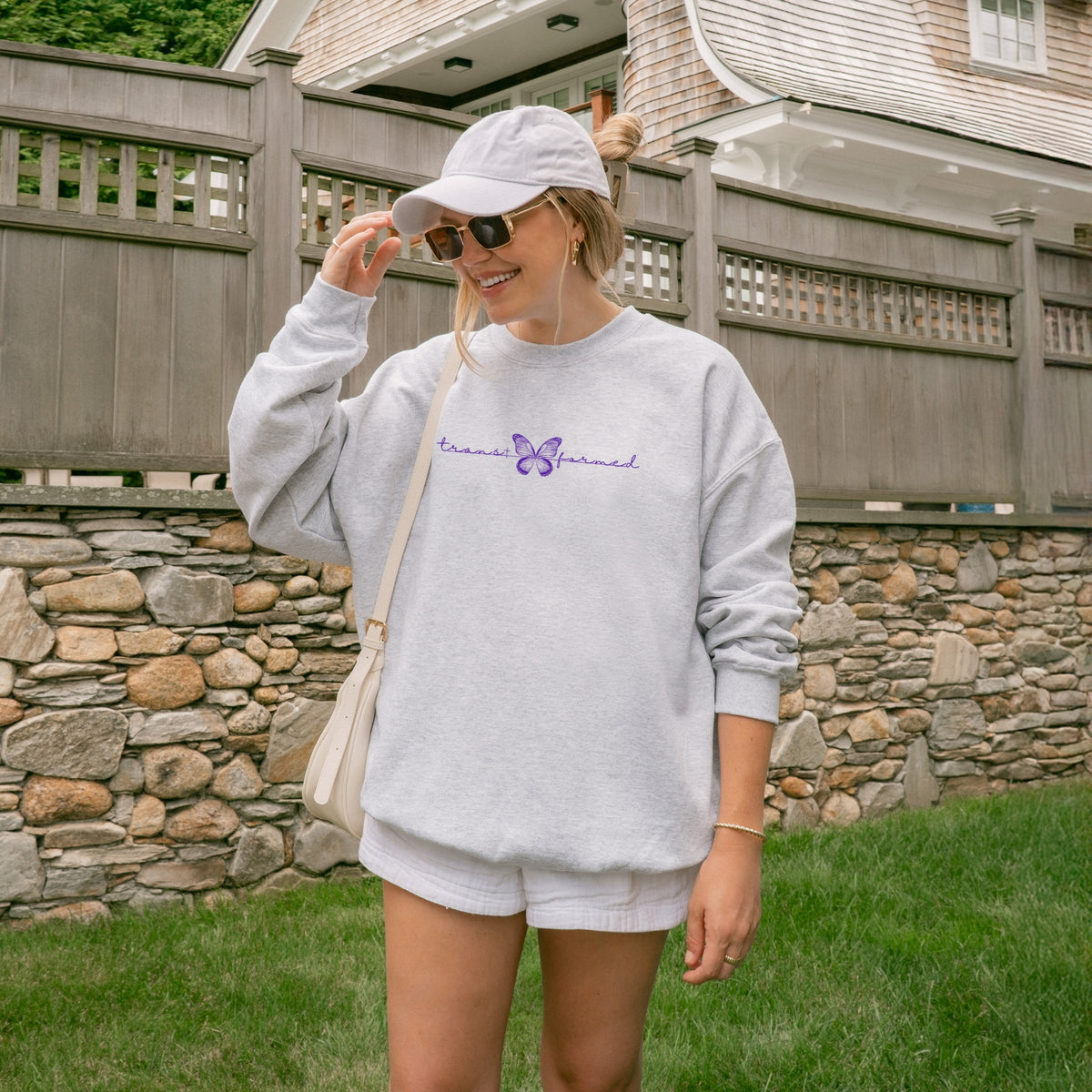 Woman wearing a gray sweatshirt with a butterfly, white shorts, and a cap, standing in front of a stone wall.