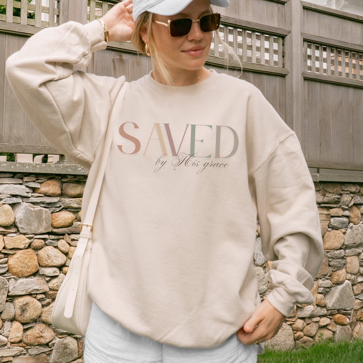 Person wearing a beige sweatshirt with 'SAVED by His grace' text, standing in front of a stone wall.