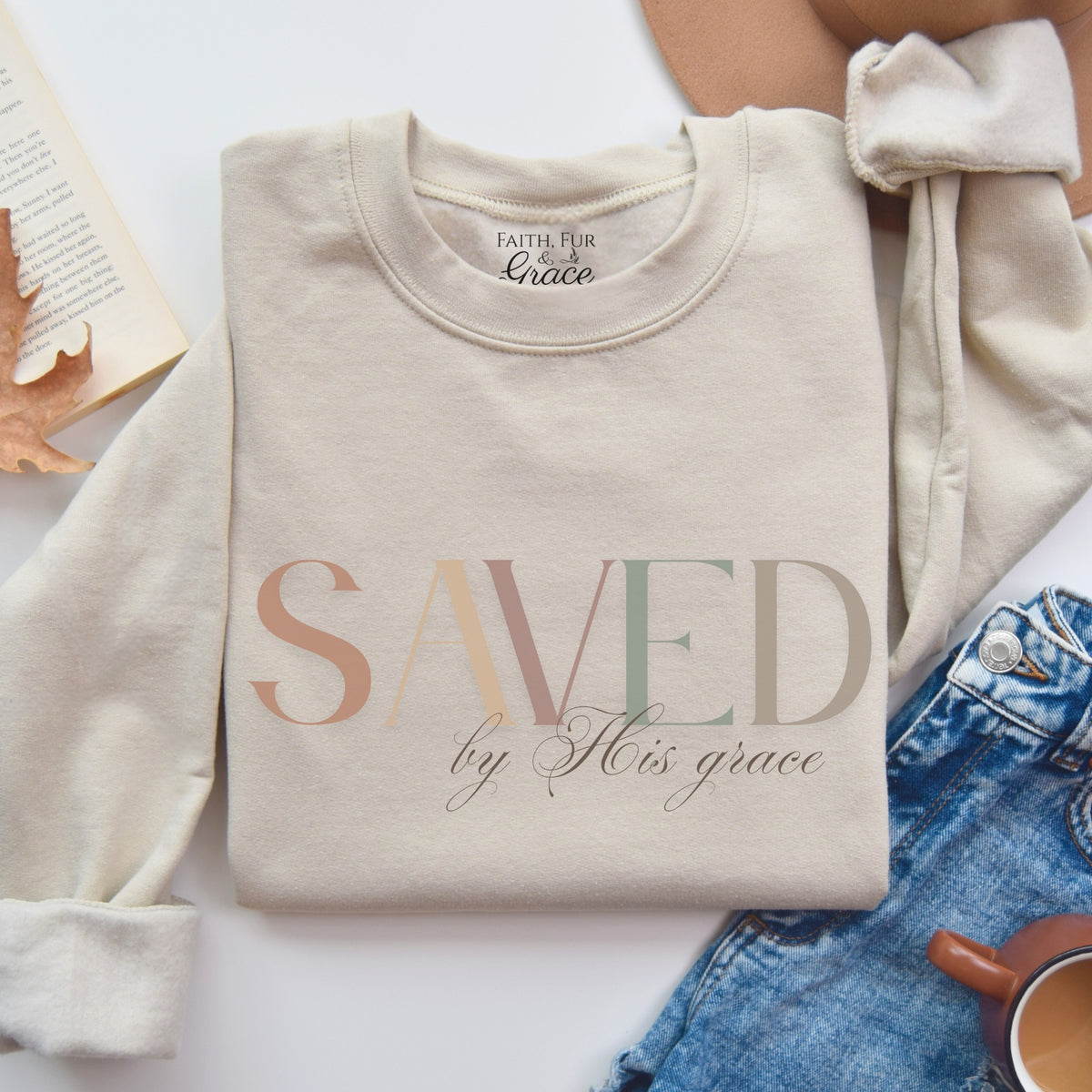Beige sweatshirt with 'SAVED by His grace' text on a white surface with denim jeans and a book.