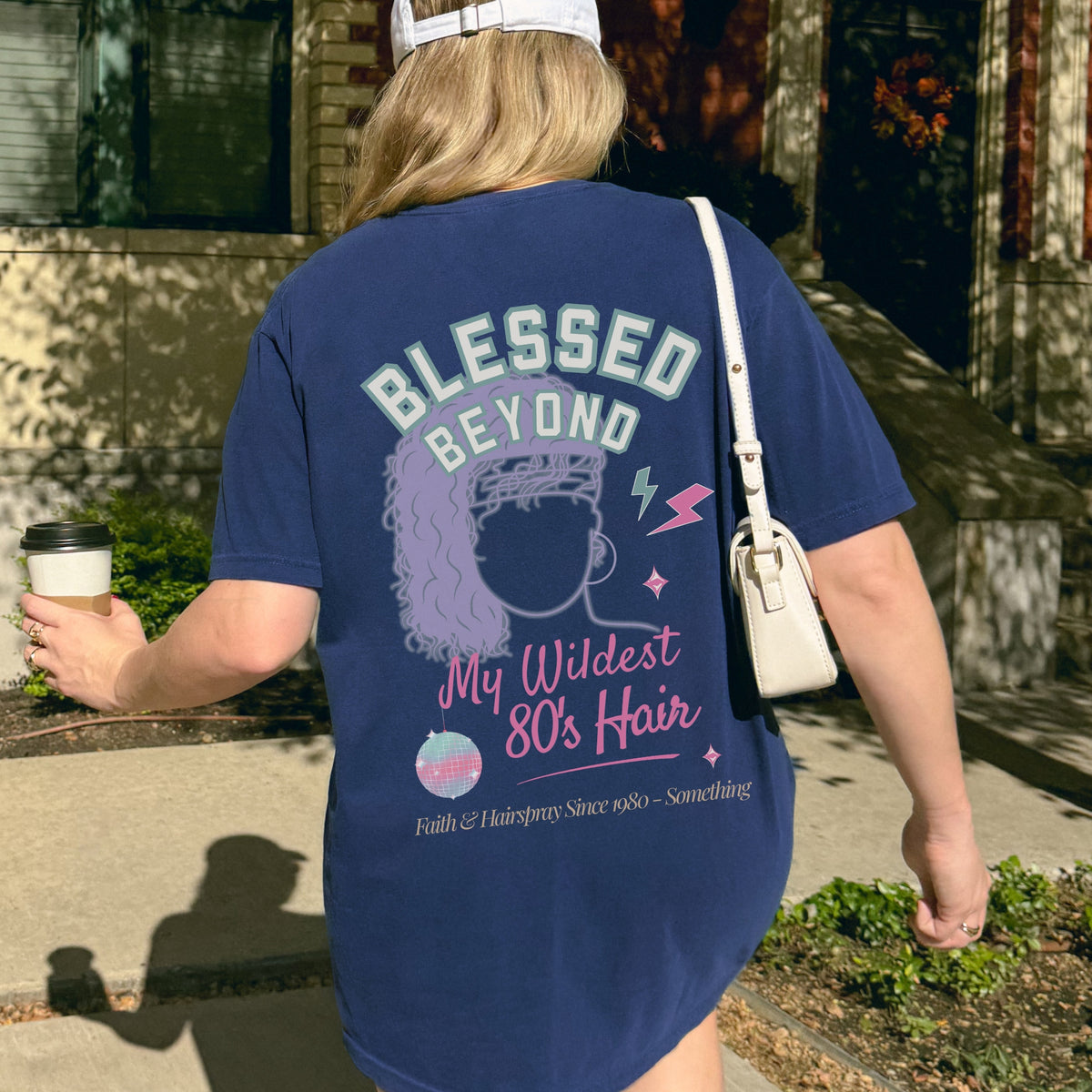 Person wearing a blue t-shirt with a graphic and text, holding a coffee cup outdoors.