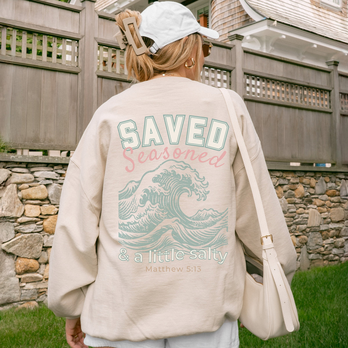 Person wearing a beige sweatshirt with a wave design and text, standing in front of a stone wall.