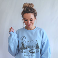 Woman wearing a light blue sweatshirt with a nature design and 'Peace Like a River' text.