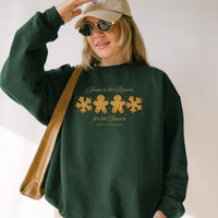 Person wearing a green sweatshirt with a Christmas design and text, holding a beige cap and sunglasses.