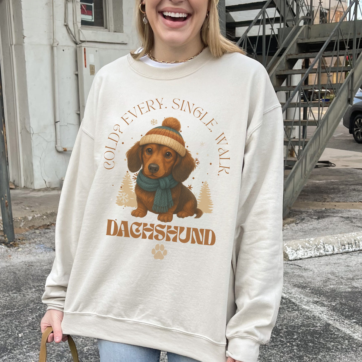 Person wearing a beige sweatshirt with a dachshund graphic and text outdoors.