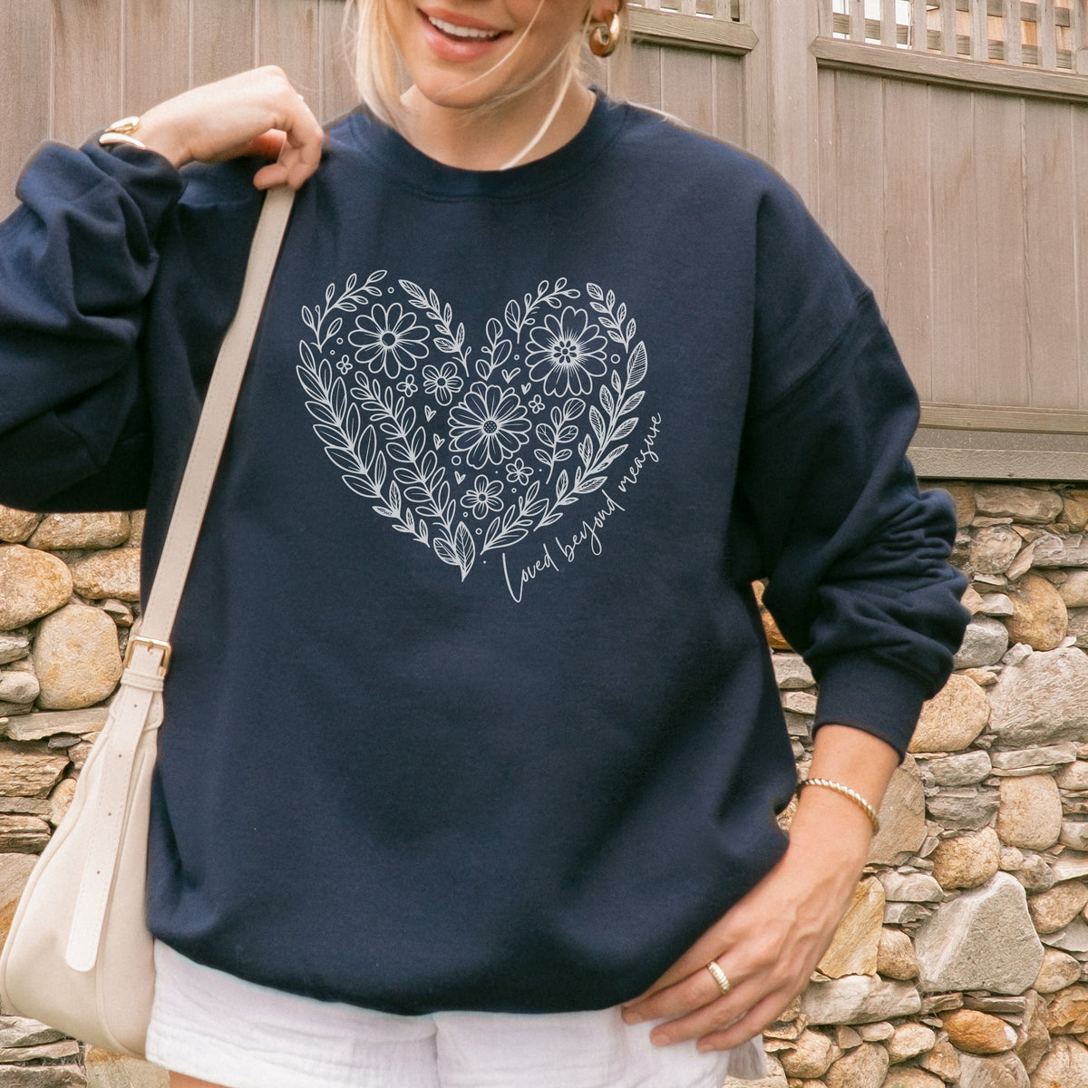 Person wearing a navy blue sweatshirt with floral heart design and text, standing against a stone wall.