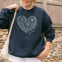 Person wearing a navy blue sweatshirt with floral heart design and text, standing against a stone wall.