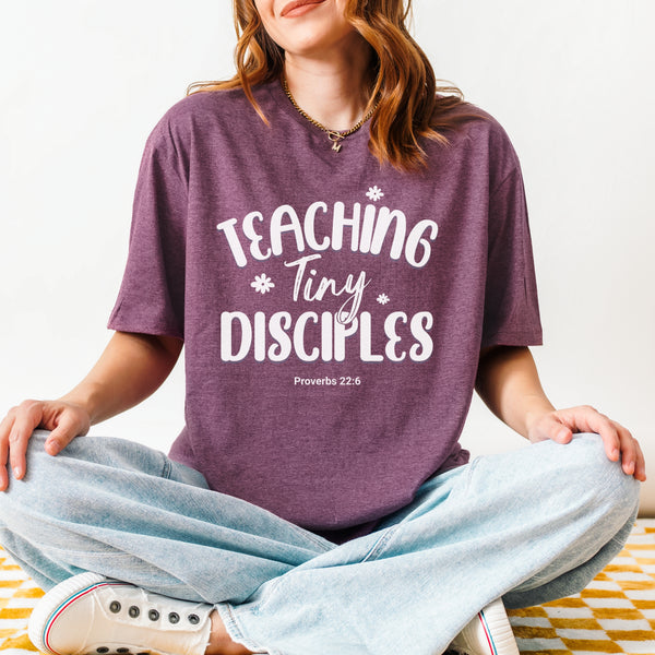 Person wearing a purple t-shirt with 'Teaching Tiny Disciples' text, sitting on a checkered floor.
