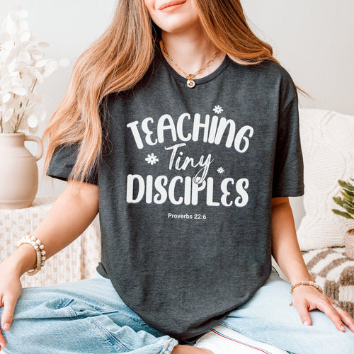 Person wearing a dark gray t-shirt with 'Teaching Tiny Disciples' text, sitting indoors.