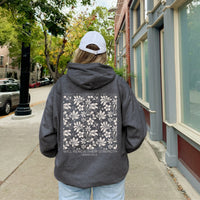 Person wearing a dark hoodie with a floral design and text on the back, walking down a street.
