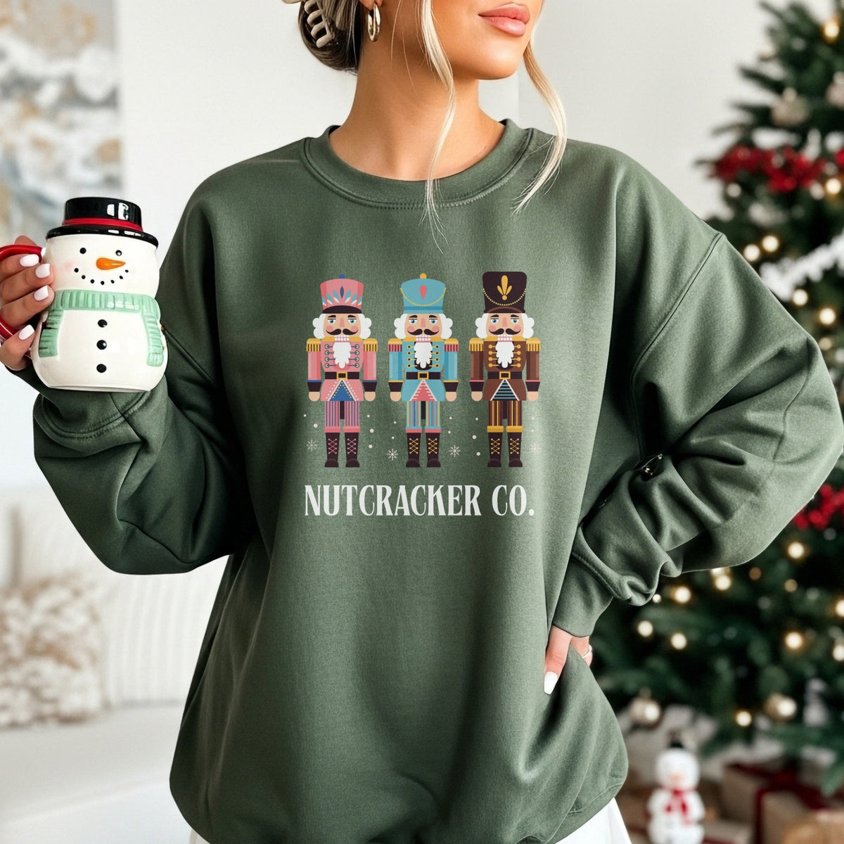 Person wearing a green sweatshirt with Nutcracker design and 'Nutcracker Co.' text, holding a mug in front of a Christmas tree.