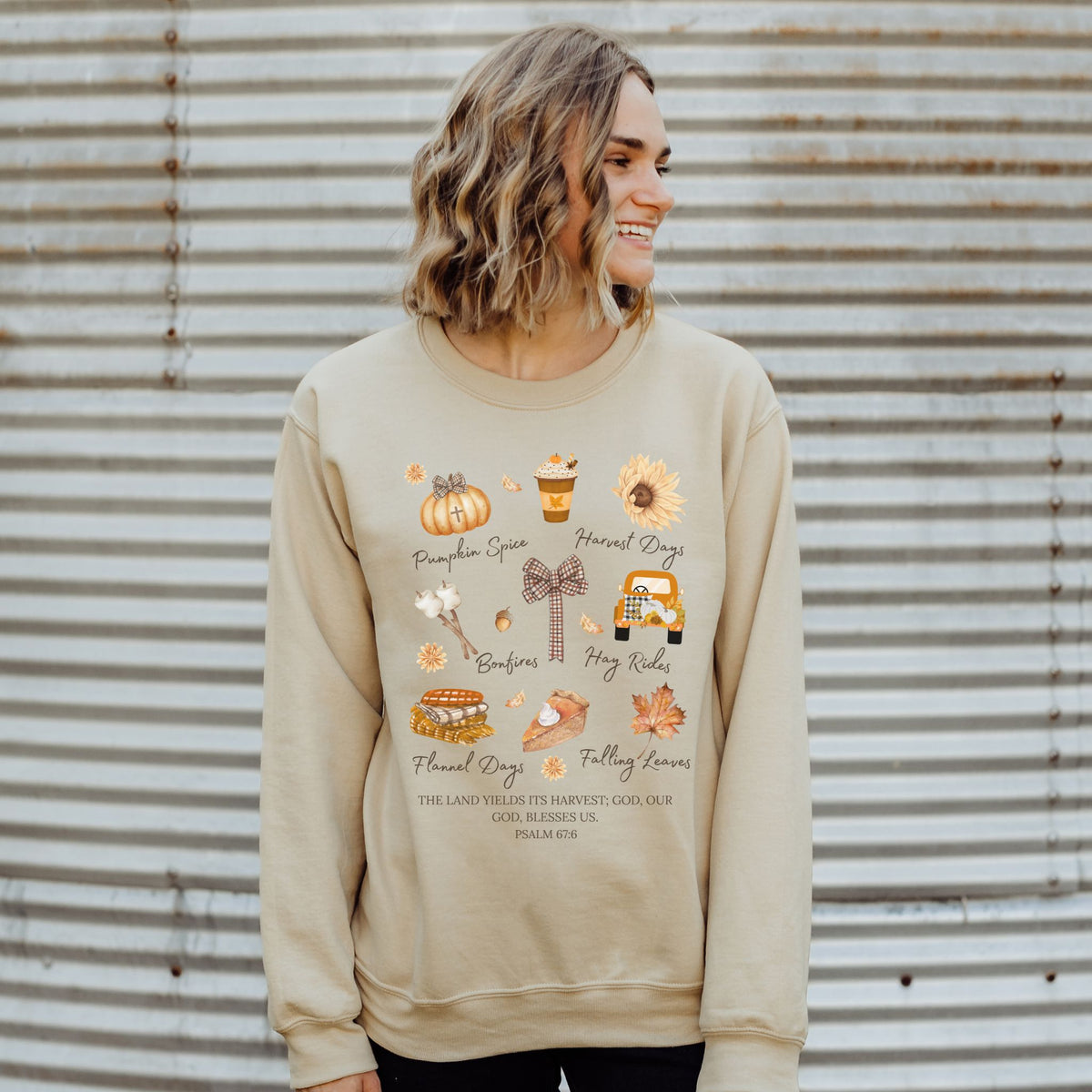 Woman wearing a beige sweatshirt with autumn-themed graphics against a corrugated metal background