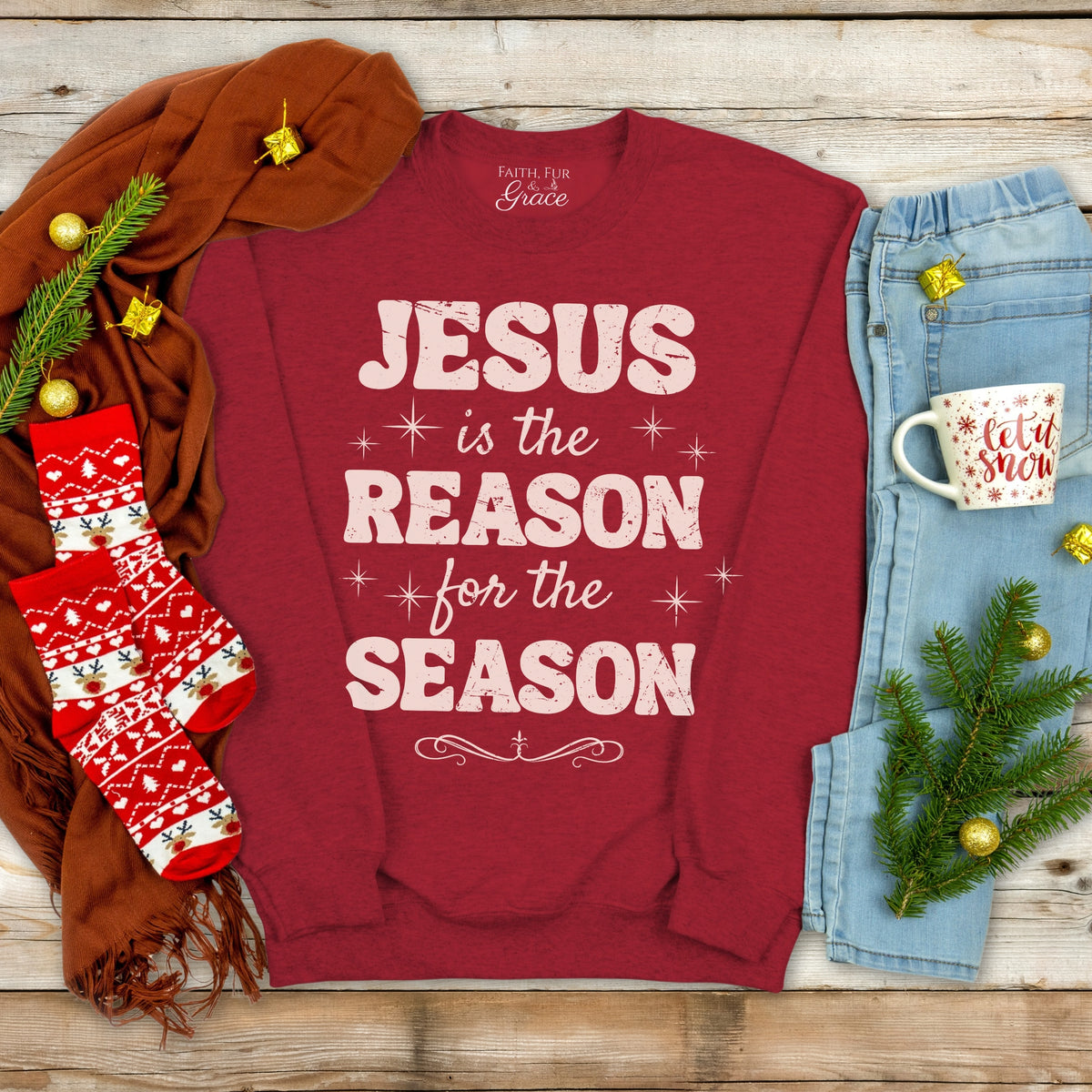 Red sweatshirt with 'Jesus is the Reason for the Season' text, surrounded by Christmas decor on a wooden surface.