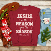 Red sweatshirt with 'Jesus is the Reason for the Season' text, surrounded by Christmas decor on a wooden surface.