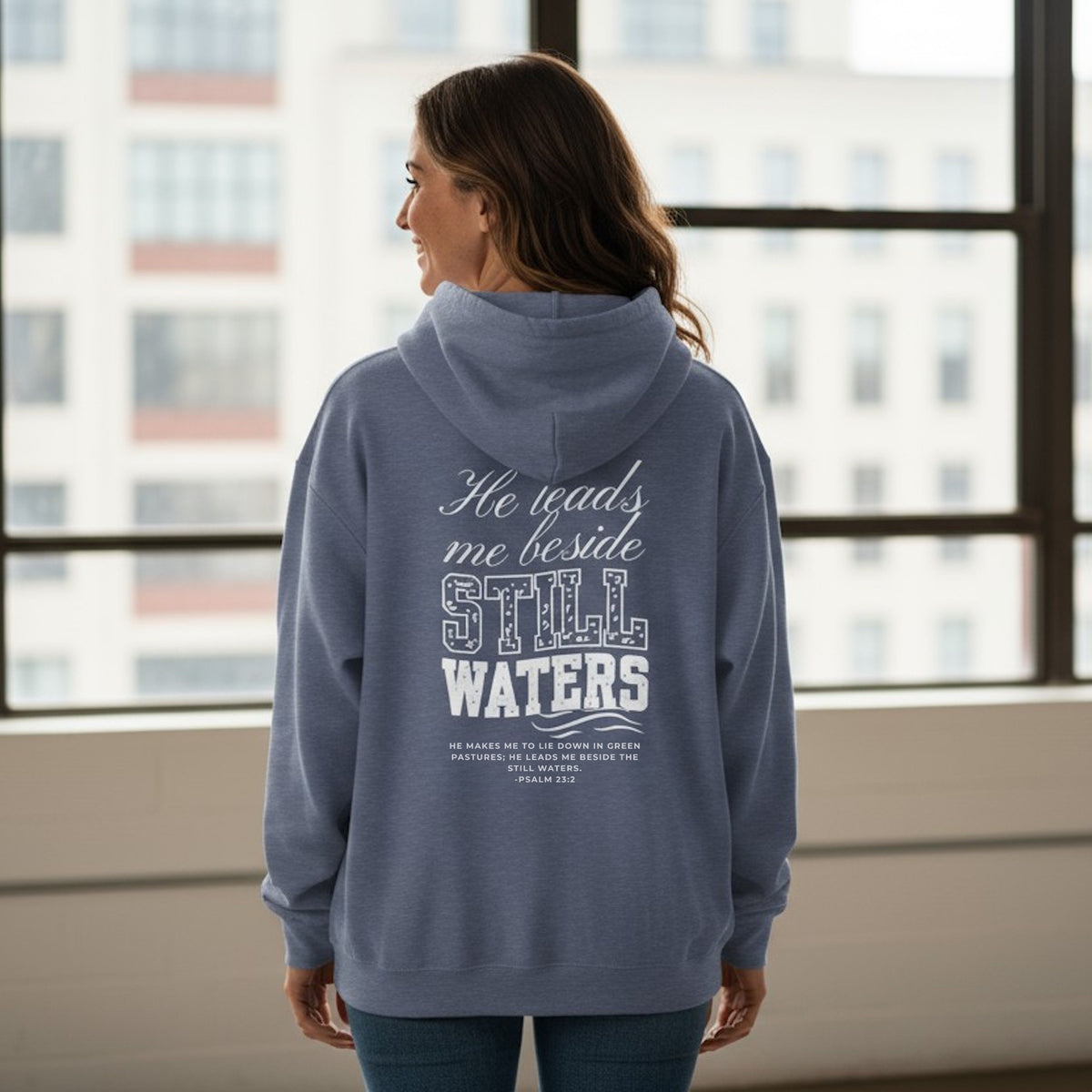 Person wearing a blue hoodie with text on the back, standing in front of a window with cityscape view.