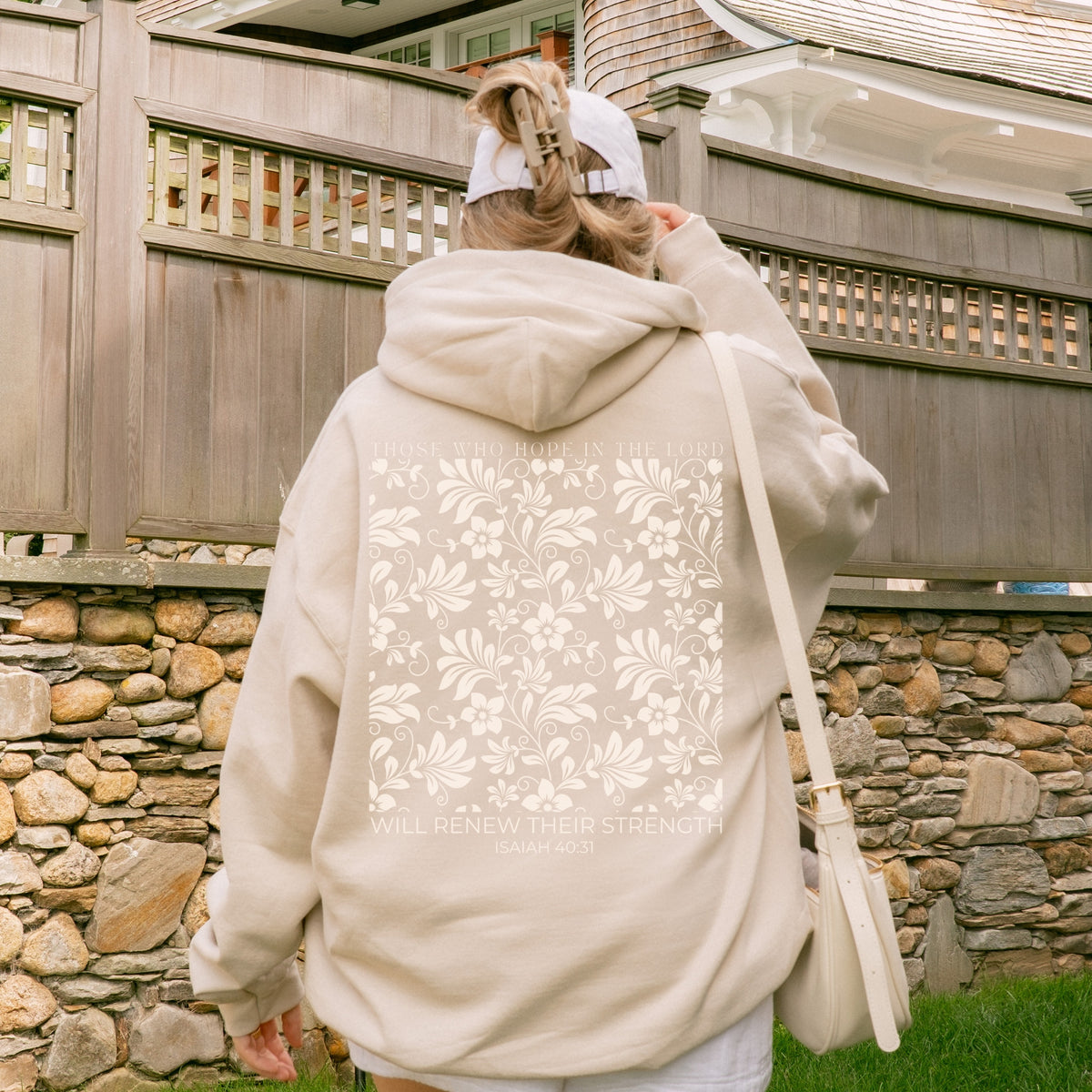 Person wearing a beige hoodie with floral patterns and text, standing outdoors near a stone wall.