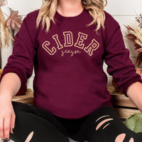 Person wearing a maroon sweatshirt with 'CIDER season' text, sitting on a wooden surface.