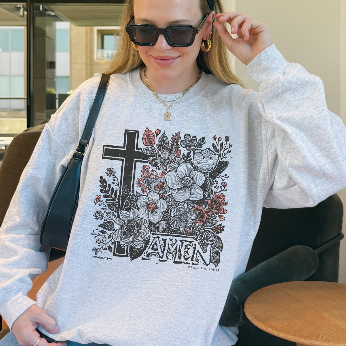 Person wearing a gray sweatshirt with floral and cross design, sitting indoors.